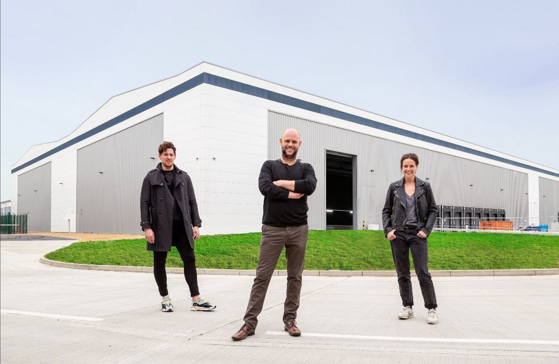 Three people stand in front of a large building and a patch of grass.