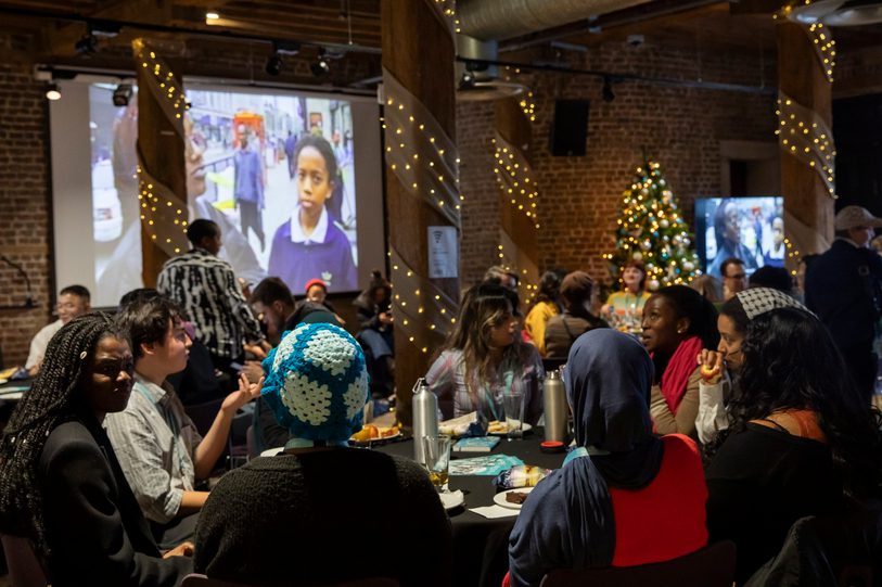 Group of people sat around tables with projector in background playing archive film