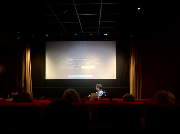 A man in a wheelchair sits in front of a cinema audience, screen says 'How many disabled peoiple are there in the UK?' with multiple choice answers - the correct answer, 14.1 million, is lit up in green.