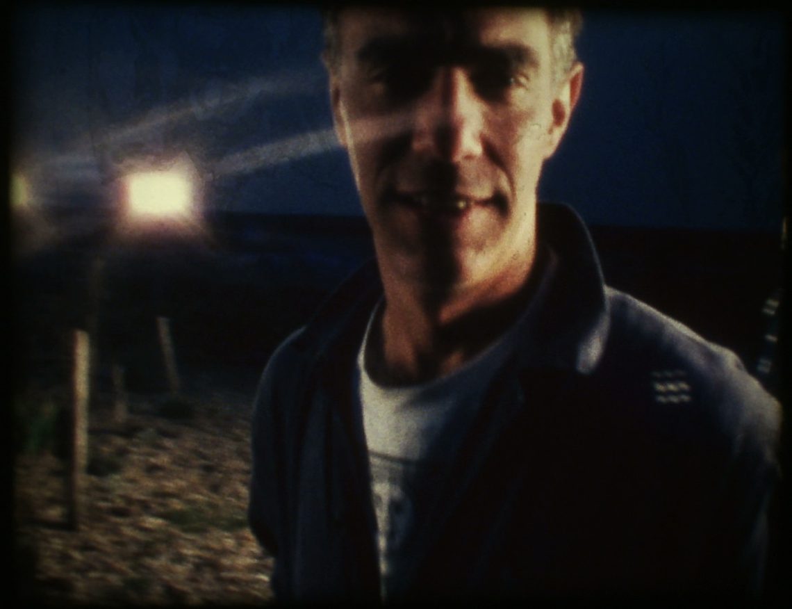 Derek Jarman looking at camera on the beach with a spotlight shining behind him.