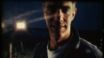 Derek Jarman looking at camera on the beach with a spotlight shining behind him.