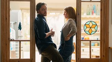 Still of Together. Two people stand in a doorframe with one looking directly at the camera