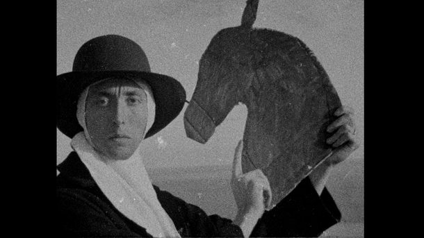 A black-and-white image depicts a figure wearing a wide-brimmed hat and wrapped head covering, holding a carved wooden horse’s head. The subject looks directly towards the viewer with a serious expression. The stark composition and historical aesthetic evoke ritual, performance, or folklore.