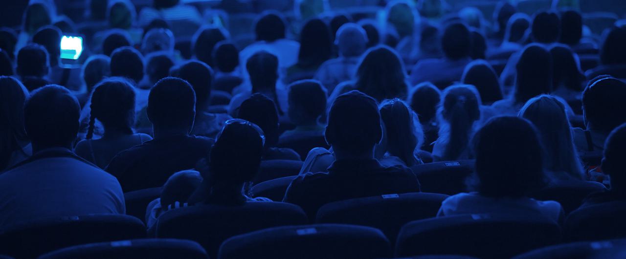 An audience in the dark in a cinema, shot from behind.